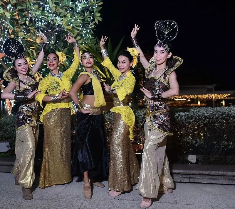 Five cultural dancers in yellow kebaya, gold sequinned skirts and tall black headdresses posing under fairy lights at an evening event in Singapore