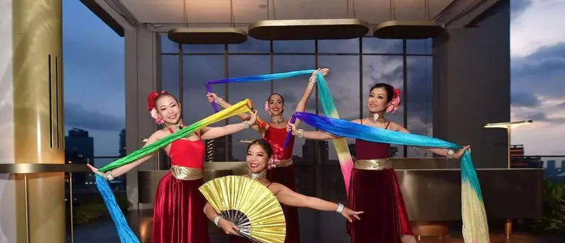 Four dancers in red velvet costumes holding long rainbow silk ribbons and a gold fan in a rooftop venue at dusk